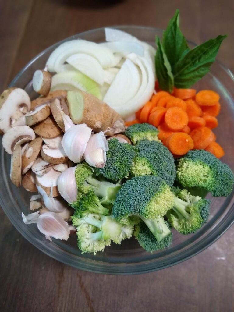 A clear bowl of fresh, crisp veggies including carrots, broccoli, mushrooms, onions, garlic, ginger and basil